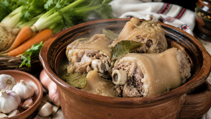 Picioare de porc fierte într-un vas tradițional, alături de usturoi și legume.
